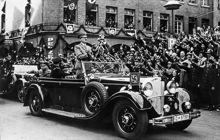 Adolf Hitler is standing in his open car while driving through Cologne